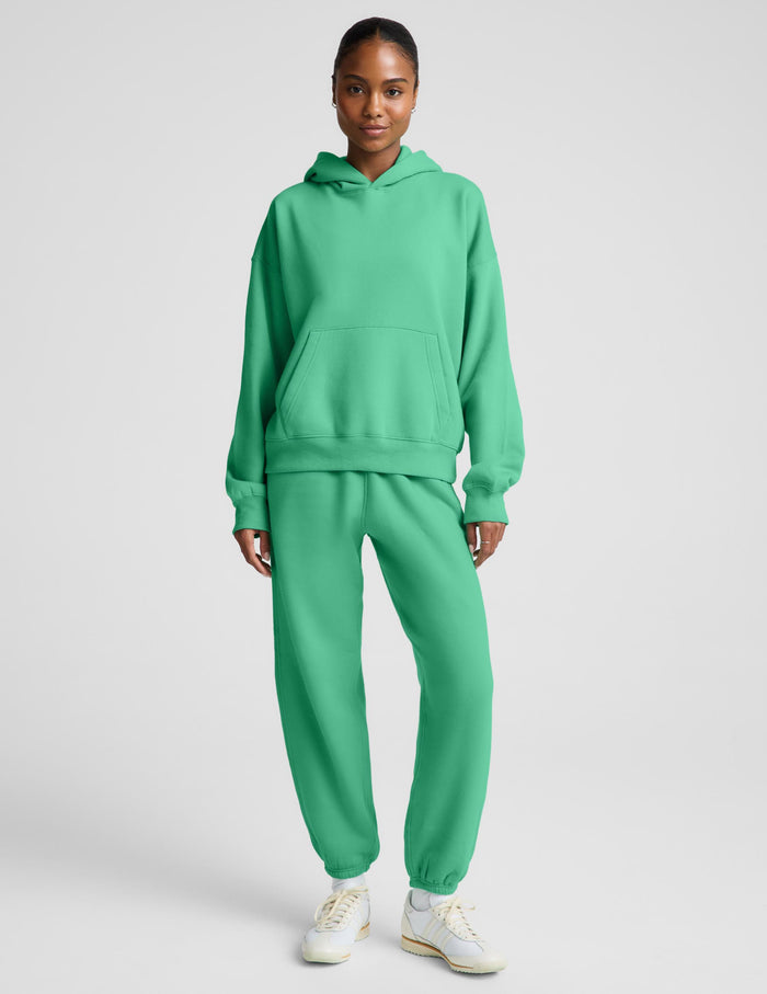 A woman wearing a bright green pullover hoodie and matching sweatpants, photographed against a plain light gray studio background.