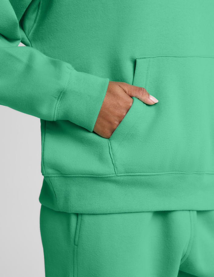 A woman wearing a bright green pullover hoodie and matching sweatpants, photographed against a plain light gray studio background.
