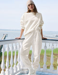 A woman wearing a matching cream-colored lounge set consisting of a long-sleeve sweatshirt and jogger pants, standing on a white wooden deck with a scenic ocean background.