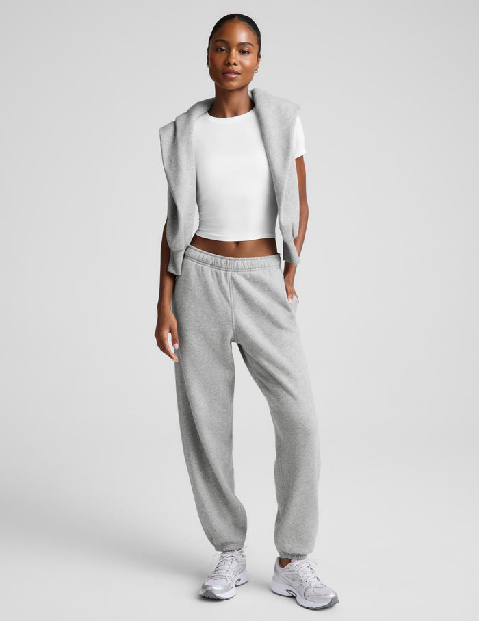 A model wearing a light gray sleeveless cropped jacket over a white crop top, paired with matching light gray sweatpants and white sneakers, set against a plain light gray studio background.