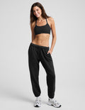 A woman modeling a black sports bra and black jogger sweatpants with white sneakers, standing against a light gray studio background.