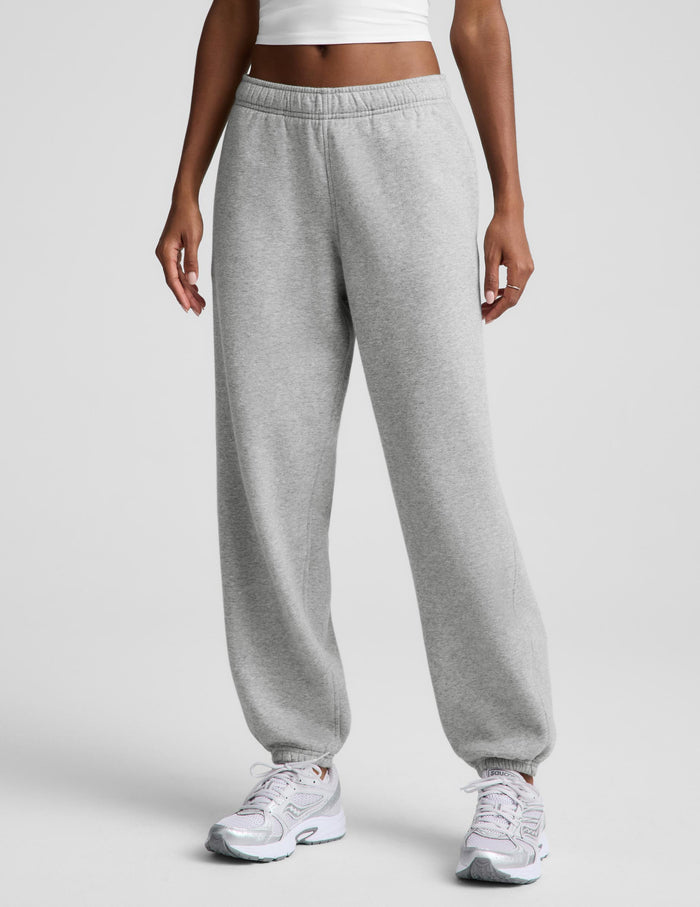 A person wearing light heather gray sweatpants with an elastic waistband and cuffs, paired with white sneakers, shown from the waist down against a light gray studio background.