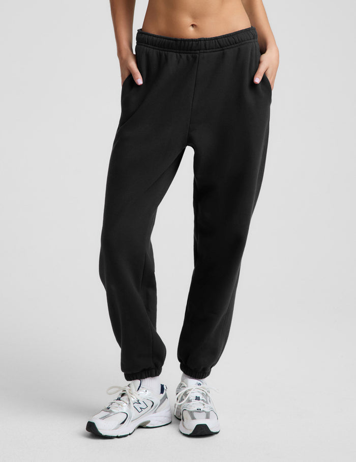 A woman modeling a black sports bra and black jogger sweatpants with white sneakers, standing against a light gray studio background.