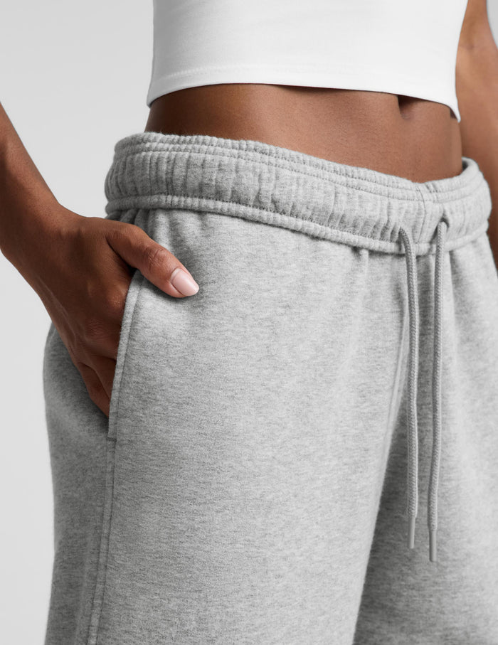 Close-up of a person wearing light heather gray sweatpants with an elastic drawstring waistband, shown with one hand in the pocket against a plain light gray background.