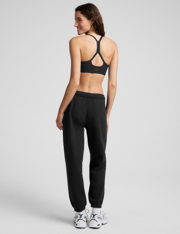 A woman modeling a black sports bra and black jogger sweatpants with white sneakers, standing against a light gray studio background.