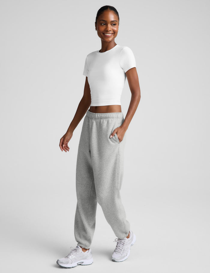 A woman modeling a white short-sleeve crop top and light gray jogger sweatpants with white sneakers, standing against a light gray studio background.