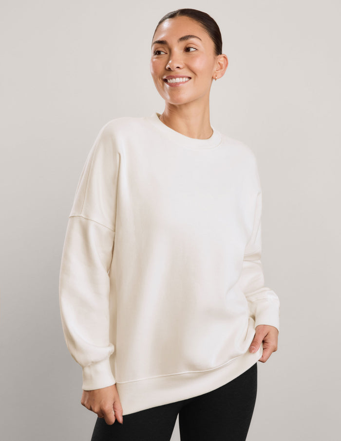 A woman wearing an oversized cream-colored crewneck sweatshirt and black leggings, against a neutral light gray studio background.