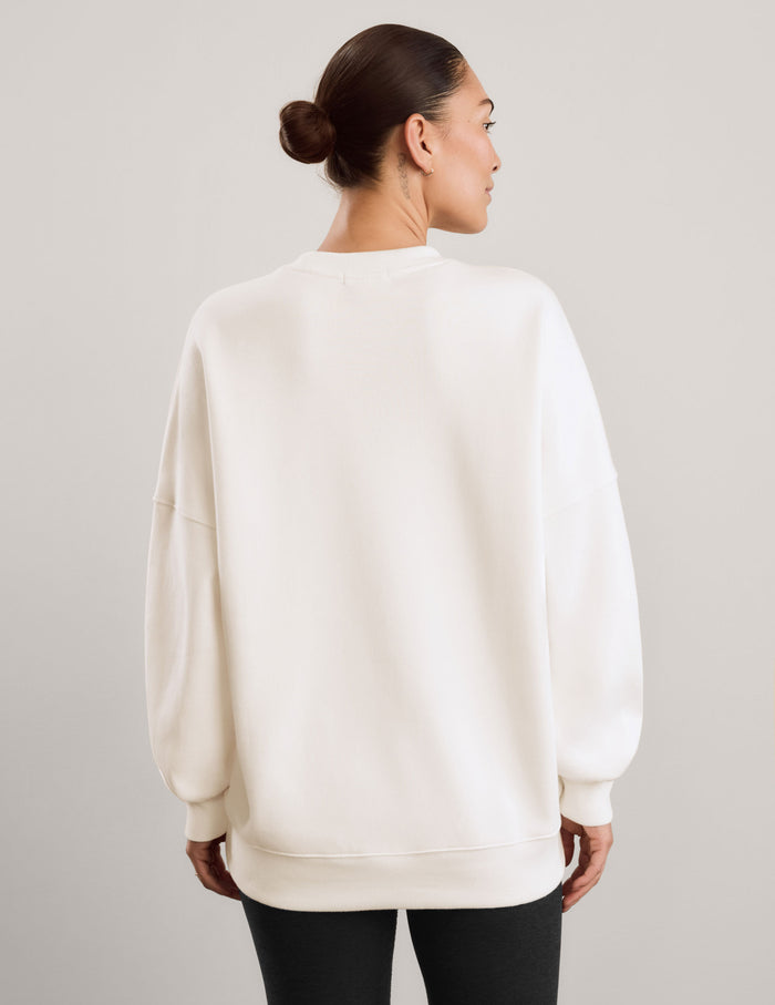 A woman wearing an oversized cream-colored crewneck sweatshirt and black leggings, against a neutral light gray studio background.