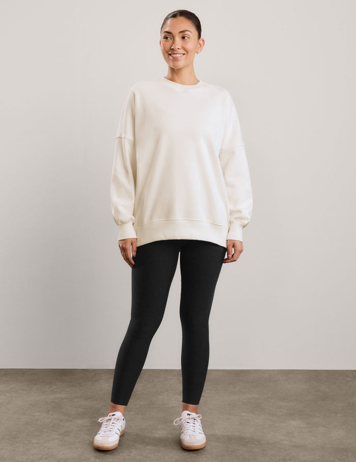 A woman wearing an oversized cream-colored crewneck sweatshirt and black leggings, against a neutral light gray studio background.
