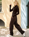 A woman modeling a dark brown long-sleeve zip-front jacket with wide-leg pants, styled with white sneakers, set against a sunlit beige stone wall.