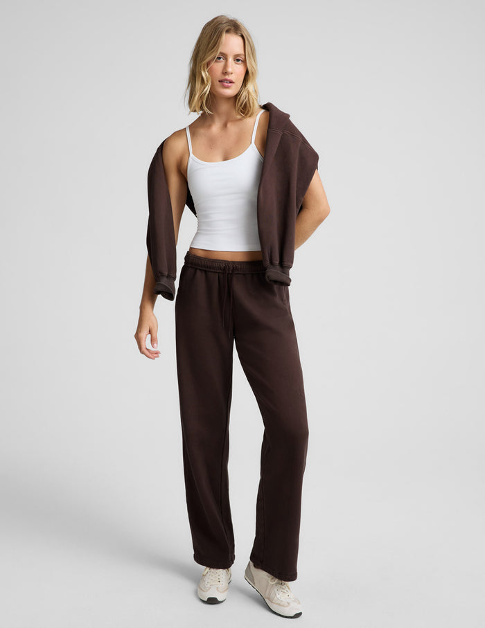 Woman wearing a white tank top and brown straight leg sweatpants with a brown sweatshirt around her shoulders against a plain background.