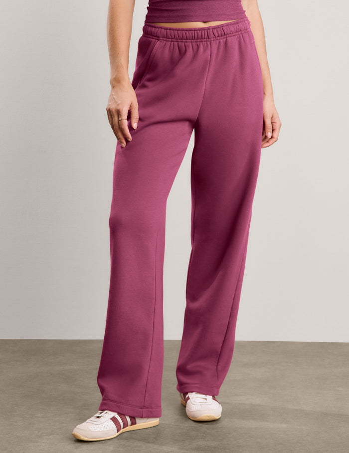 A woman modeling a matching two-piece lounge set consisting of a tank top and wide-leg sweatpants in a solid deep berry/plum color, paired with white sneakers, against a neutral studio backdrop.