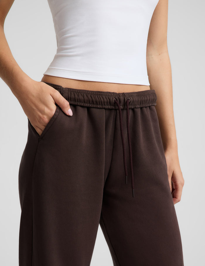 Woman wearing a white tank top and brown straight leg sweatpants against a plain background.