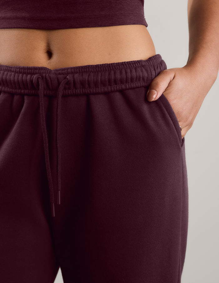 Close-up of a person wearing dark burgundy sweatpants with an elastic drawstring waistband and side pockets, paired with a matching dark burgundy crop top against a light gray studio background.
