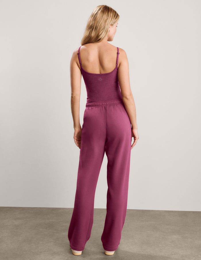 A woman modeling a matching two-piece lounge set consisting of a tank top and wide-leg sweatpants in a solid deep berry/plum color, paired with white sneakers, against a neutral studio backdrop.