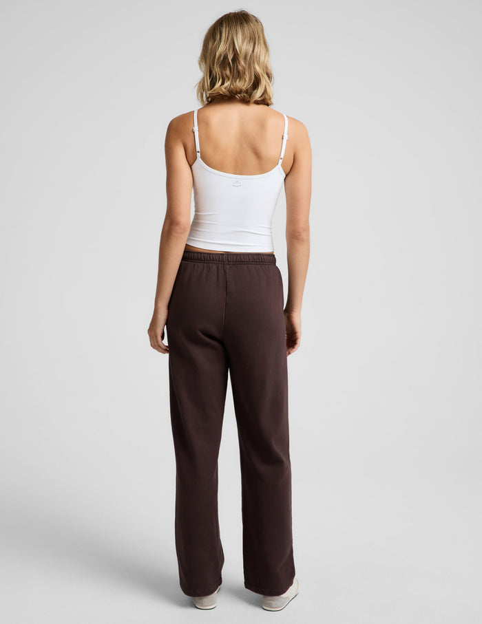 Woman wearing a white tank top and brown straight leg sweatpants against a plain background.