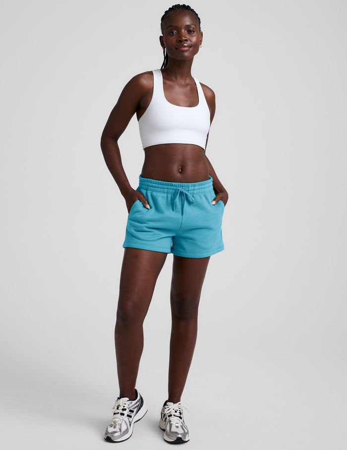 Blue LuxeFleece Sweat Shorts with drawstring waist and pockets on a model wearing a white sports bra and sneakers.