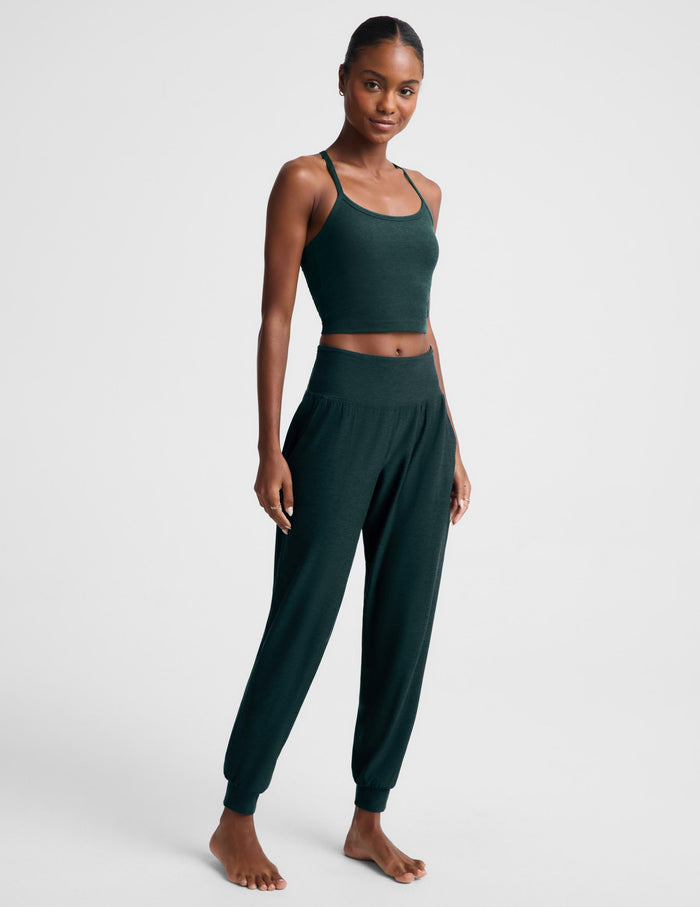 A woman modeling a dark forest green two-piece loungewear set consisting of a cropped tank top and matching jogger pants, shot against a light gray studio background.
