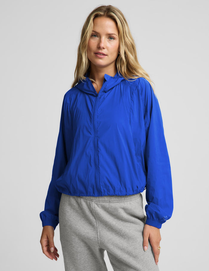 A woman wearing a bright royal blue lightweight zip-up hooded jacket and light gray sweatpants, standing against a plain light gray studio background.