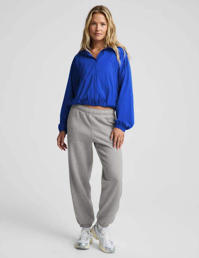 A woman modeling a bright blue cropped zip-up windbreaker jacket and light gray sweatpants against a light gray studio background.