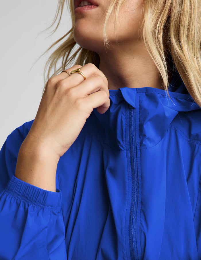 Close-up of a model wearing a bright royal blue lightweight zip-front jacket, with the model's hand adjusting the collar and zipper.