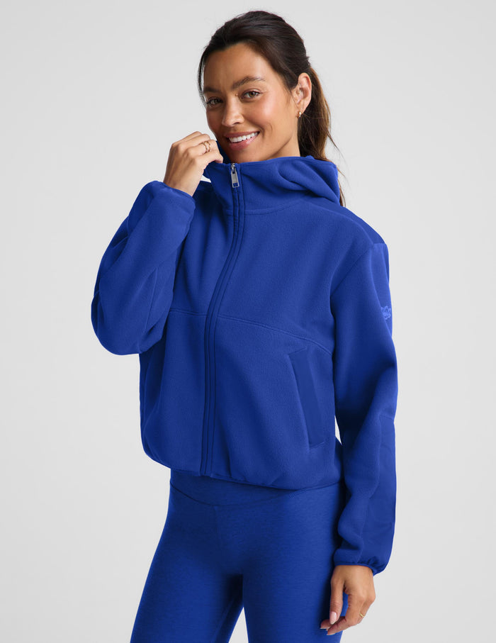 A woman wearing a bright royal blue cropped zip-up hoodie with a hood and matching blue leggings, photographed against a clean light gray studio background.