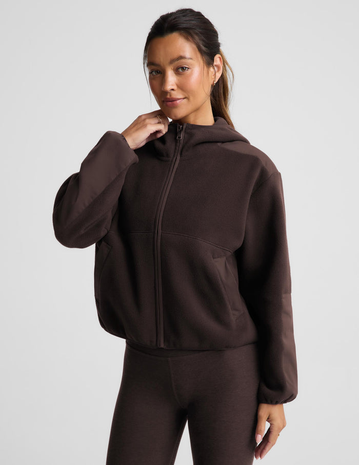 A woman modeling a dark brown cropped zip-up hoodie with a hood and matching dark brown leggings against a light gray studio background.