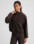 A woman modeling a dark brown cropped zip-up hoodie with a hood and matching dark brown leggings against a light gray studio background.