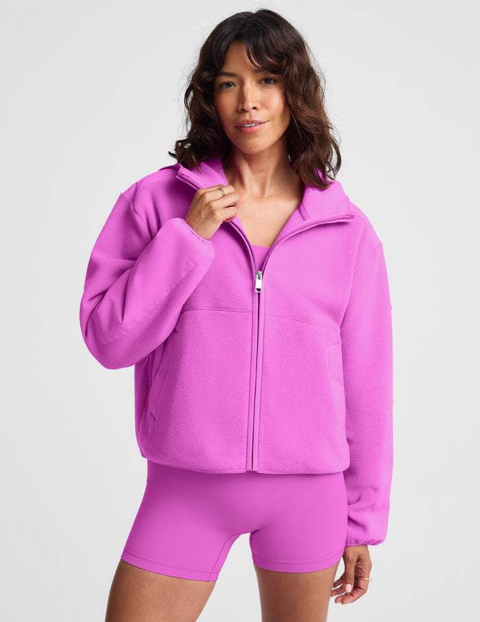 A woman modeling a bright pink zip-up fleece jacket with a stand collar, paired with matching pink athletic shorts, against a light gray studio background.