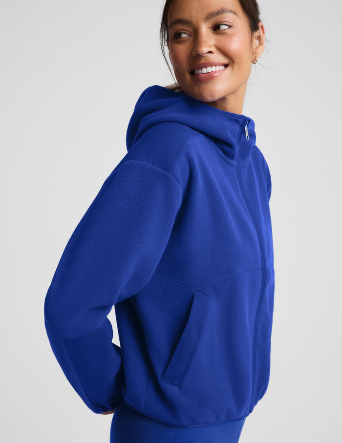 A woman wearing a bright royal blue pullover hoodie, shown from a side-back angle against a light gray studio background.