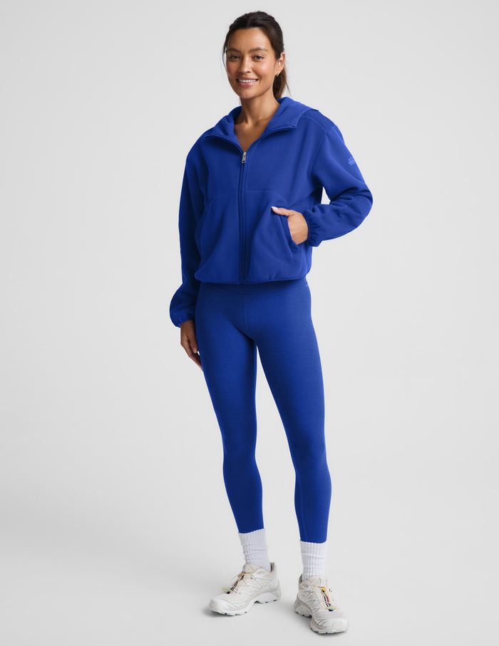 A woman modeling a matching royal blue athletic set consisting of a zip-up hoodie and high-waisted leggings, paired with white socks and white sneakers, against a light gray studio background.