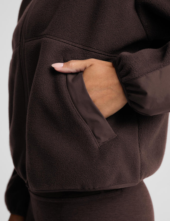 Close-up of a person wearing a dark brown fleece jacket, with one hand tucked into a side pocket. The fabric texture is clearly visible, and the background is a clean, neutral light gray.