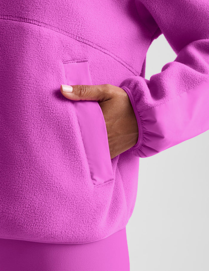 Close-up of a person wearing a bright pink fleece pullover, showing a hand inserted into a front patch pocket against a light gray background.