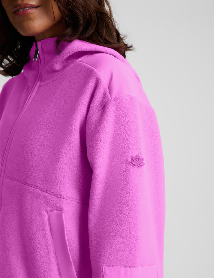 Close-up of a person wearing a bright pink hooded sweatshirt with a partial zipper, pocket, and a small tonal embroidered logo on the upper sleeve, against a light gray studio background.