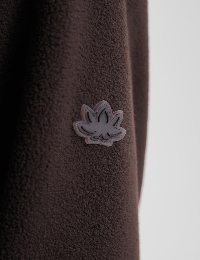 Close-up of a dark brown textured fleece garment featuring a small gray and white embroidered leaf-shaped patch on the chest, set against a light gray background.