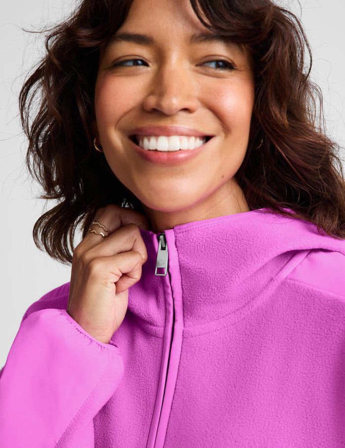 A close-up of a smiling woman wearing a bright pink fleece zip-up hoodie, adjusting the zipper with her hand against a light gray background.