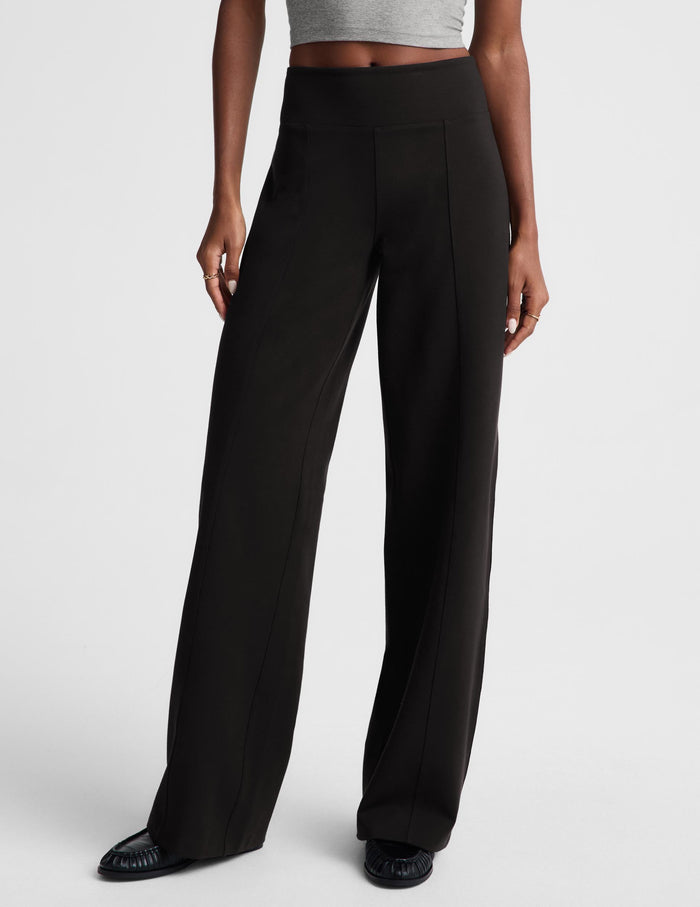 A model stands against a light gray studio background wearing a light gray crewneck sweatshirt and black wide-leg trousers.
