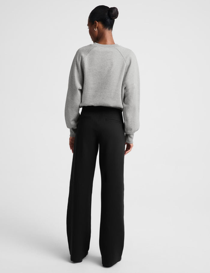 A model stands against a light gray studio background wearing a light gray crewneck sweatshirt and black wide-leg trousers.