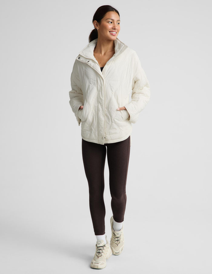 A woman modeling a cream quilted puffer jacket, paired with dark brown leggings and beige sneakers, against a solid light gray background.