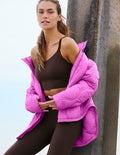 A woman wearing a bright pink puffer jacket over a brown sports bra and leggings, leaning against a concrete pillar outdoors.