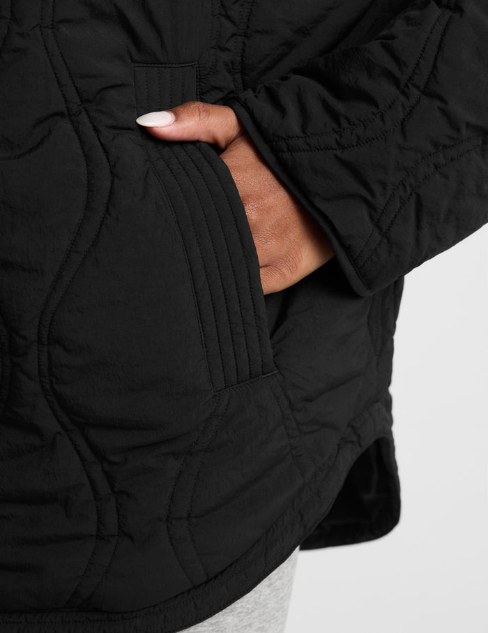 A woman wearing a black quilted zip-up jacket with a high stand-up collar, paired with light gray leggings, photographed against a plain light gray studio background.