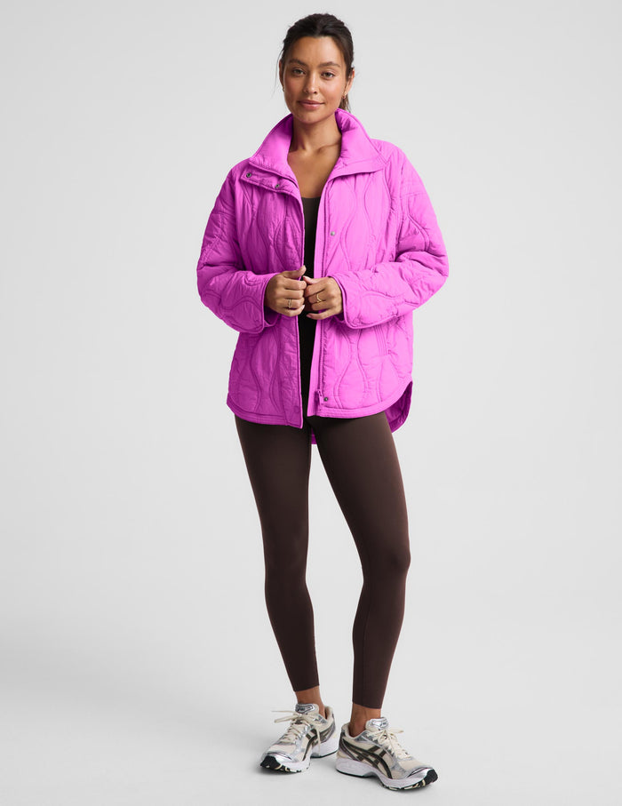A woman modeling a bright pink quilted zip-up jacket, paired with dark brown leggings and white athletic sneakers, standing against a light gray studio background.