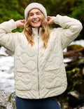 A woman outdoors wearing a cream quilted puffer jacket with a full zip and two front patch pockets, paired with a matching knit beanie and dark blue leggings. The background shows a blurred natural setting with green foliage and rocks.