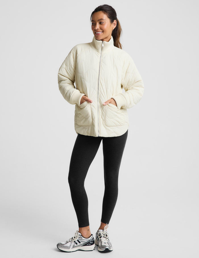 A woman modeling a cream-colored quilted zip-up jacket, paired with black leggings and silver sneakers, against a light gray studio background.