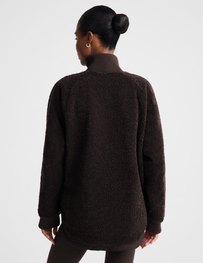 A woman shown from the back wearing a dark brown textured fleece turtleneck sweater against a light gray studio background.