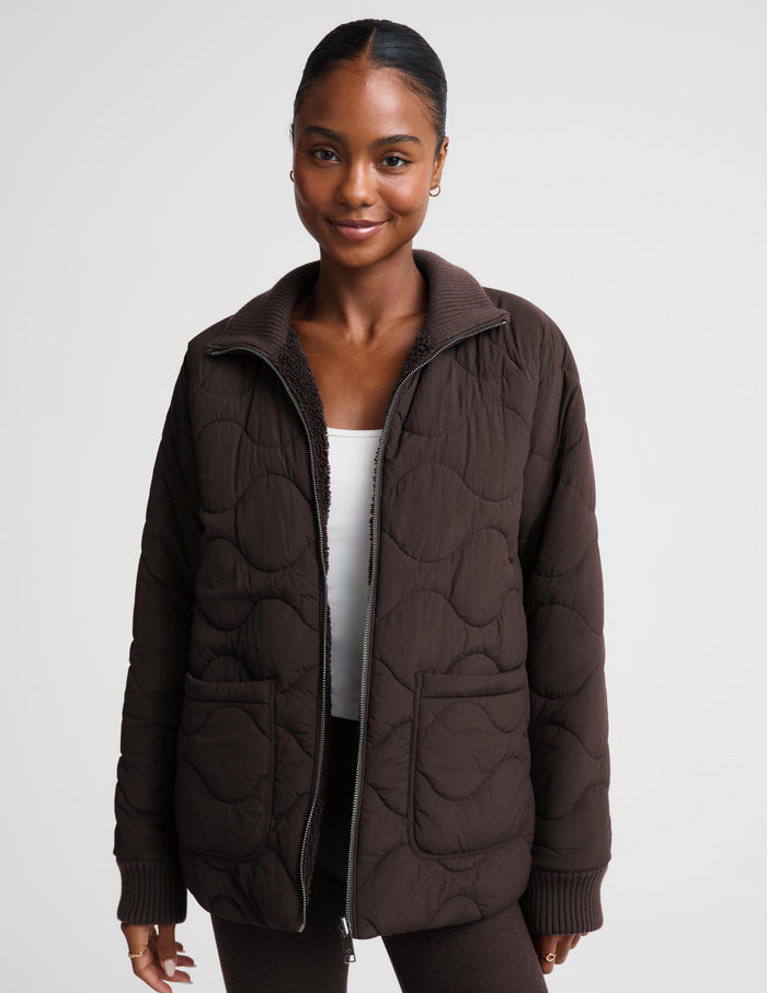 A woman wearing a dark brown quilted zip-up jacket with a ribbed collar, cuffs, and hem, layered over a white top and dark pants, photographed against a light gray studio background.