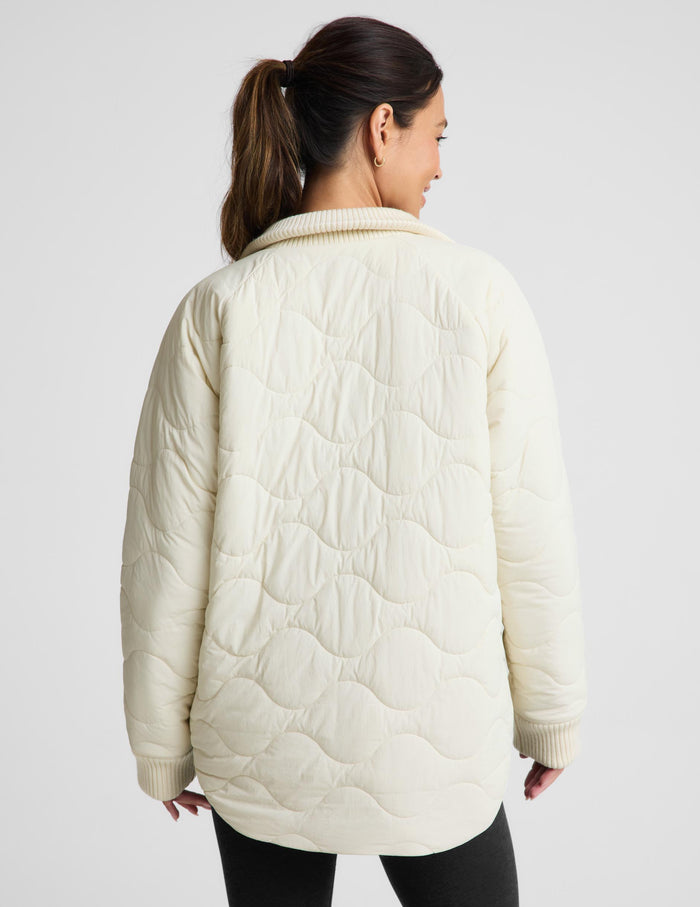 A woman shown from the back wearing a cream-colored quilted jacket with a wavy stitched pattern and ribbed collar, cuffs, and hem, paired with black pants against a light gray studio background.