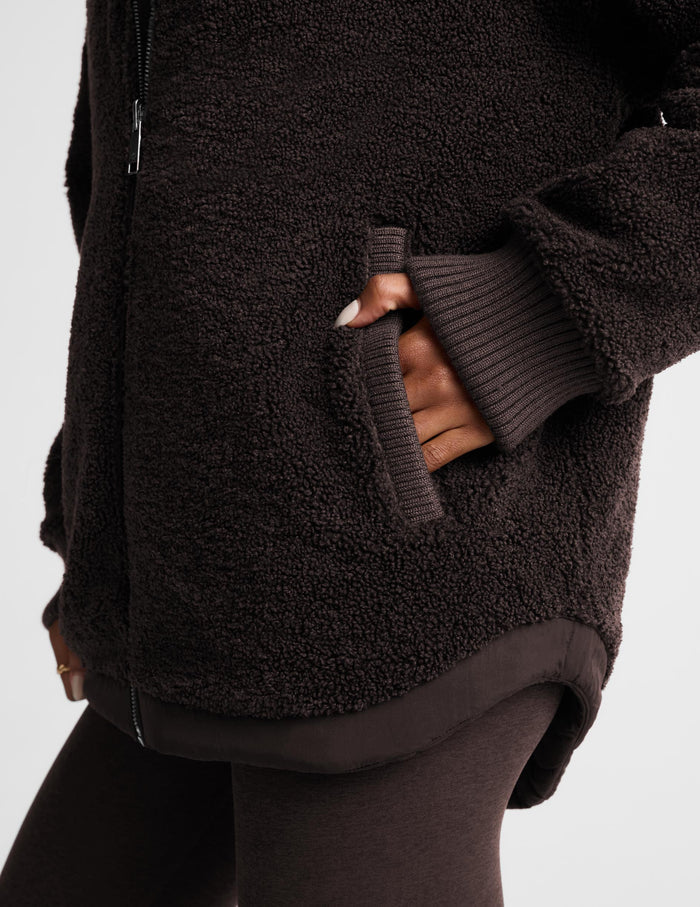 Close-up of a person wearing a dark brown sherpa fleece zip-up jacket with ribbed knit cuffs and a matching dark brown ribbed hem, paired with dark leggings against a light gray background.