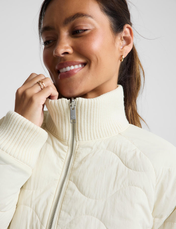A woman modeling a cream-colored quilted zip-up jacket, paired with black leggings and silver sneakers, against a light gray studio background.