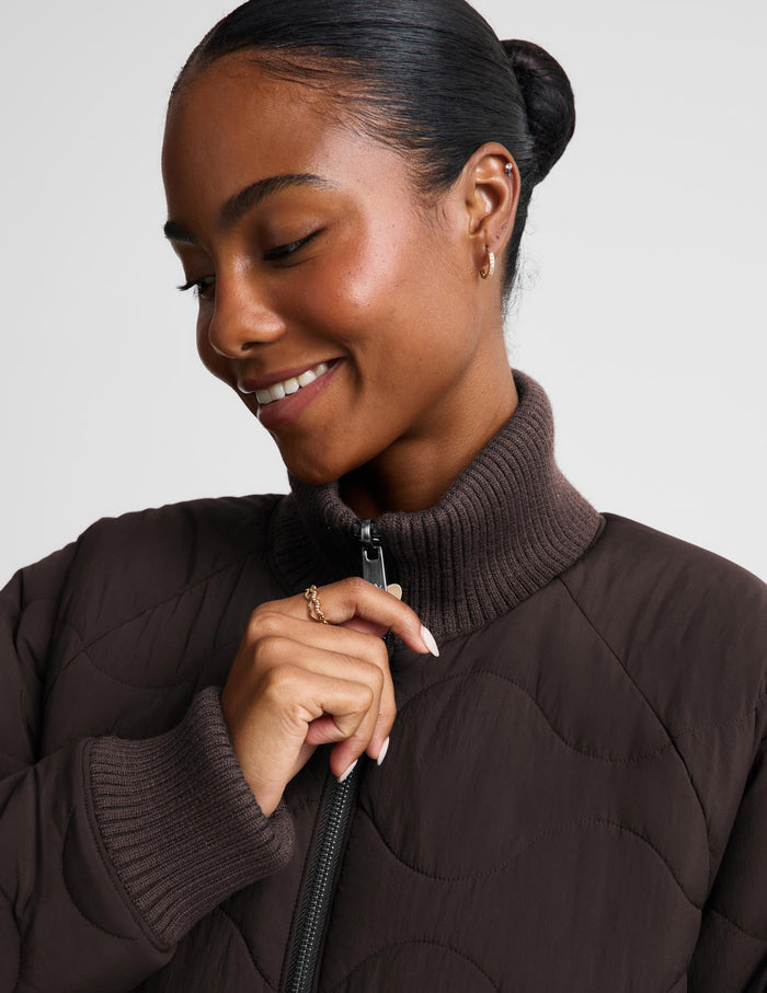 A close-up of a woman wearing a dark brown quilted puffer jacket with a ribbed mock neck and zipper, set against a plain light gray background.